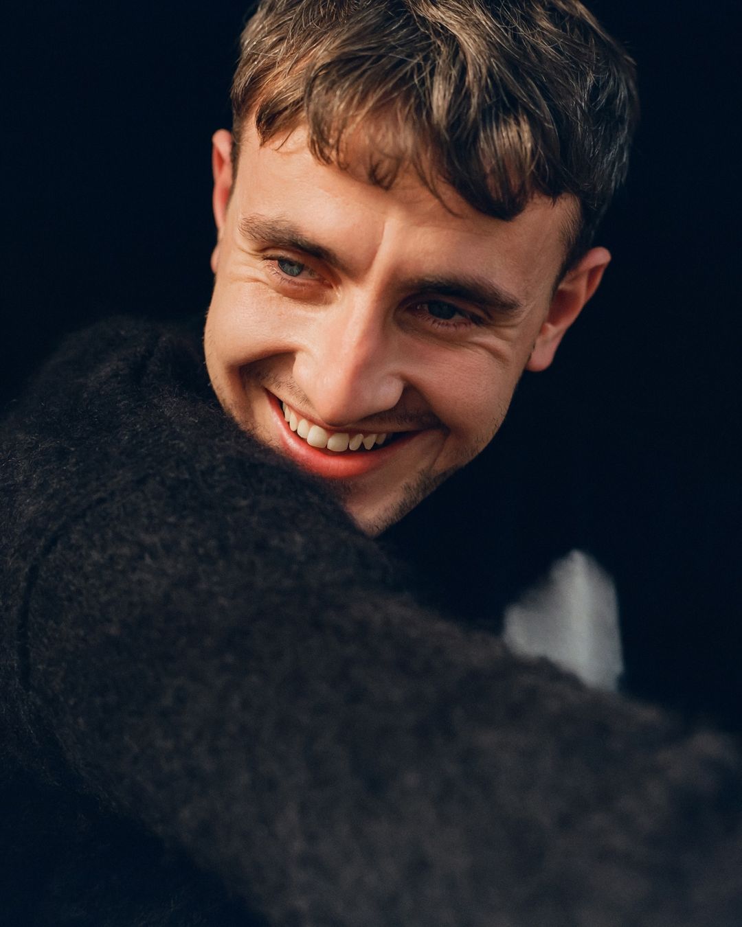 Portrait of actor Paul Mescal smiling broadly in a dark textured sweater against a plain dark background.