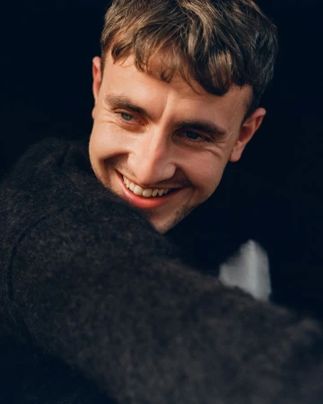 Portrait of actor Paul Mescal smiling broadly in a dark textured sweater against a plain dark background.