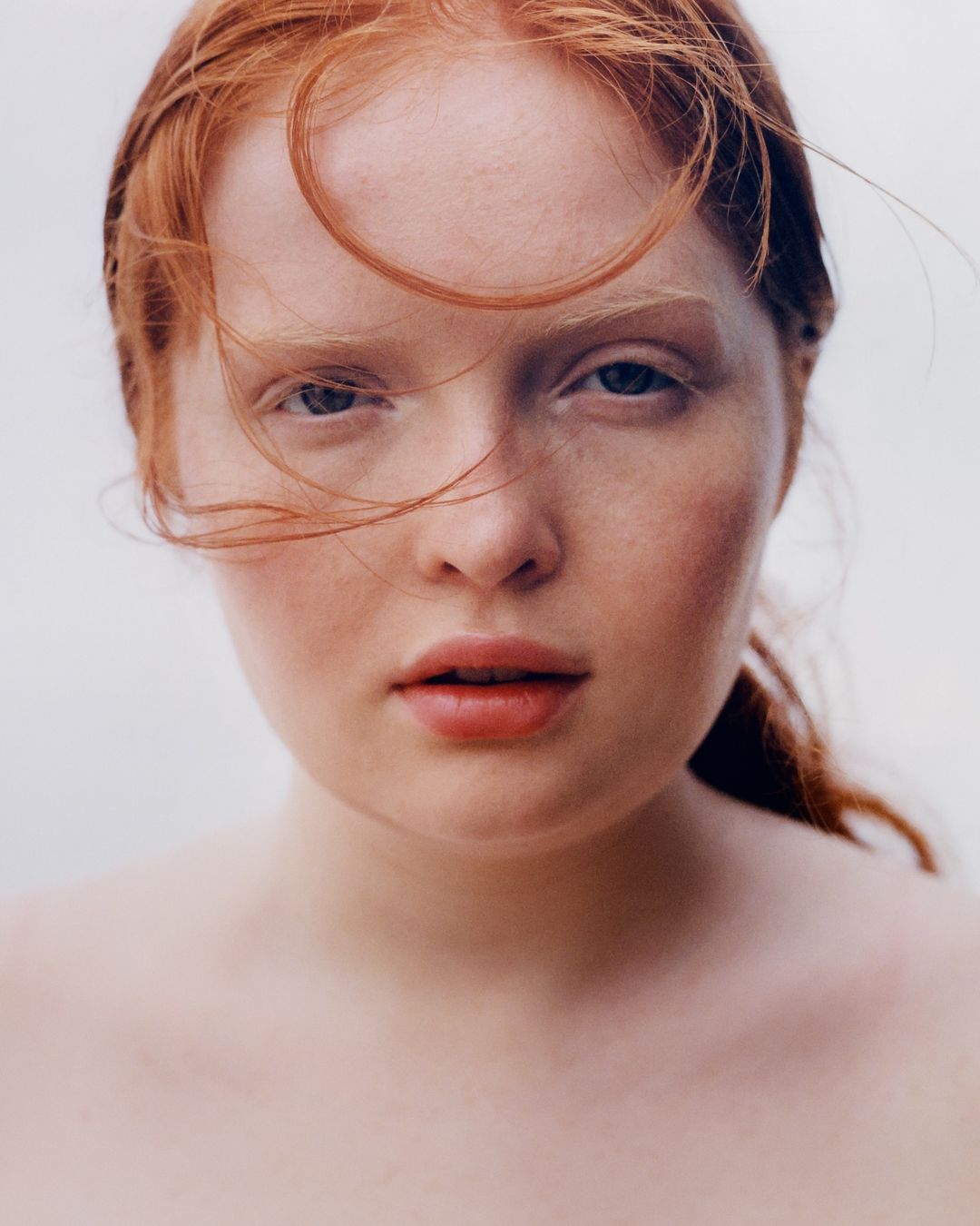 Close-up portrait of a young adult female with fair skin and striking red hair, blue eyes, and soft facial features, against a bright background.