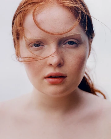 Close-up portrait of a young adult female with fair skin and striking red hair, blue eyes, and soft facial features, against a bright background.