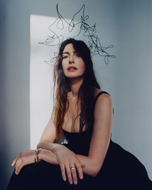 Woman in black dress poses indoors with abstract headpiece. Portrait features jewelry, indoor lighting, and fair skin tones.