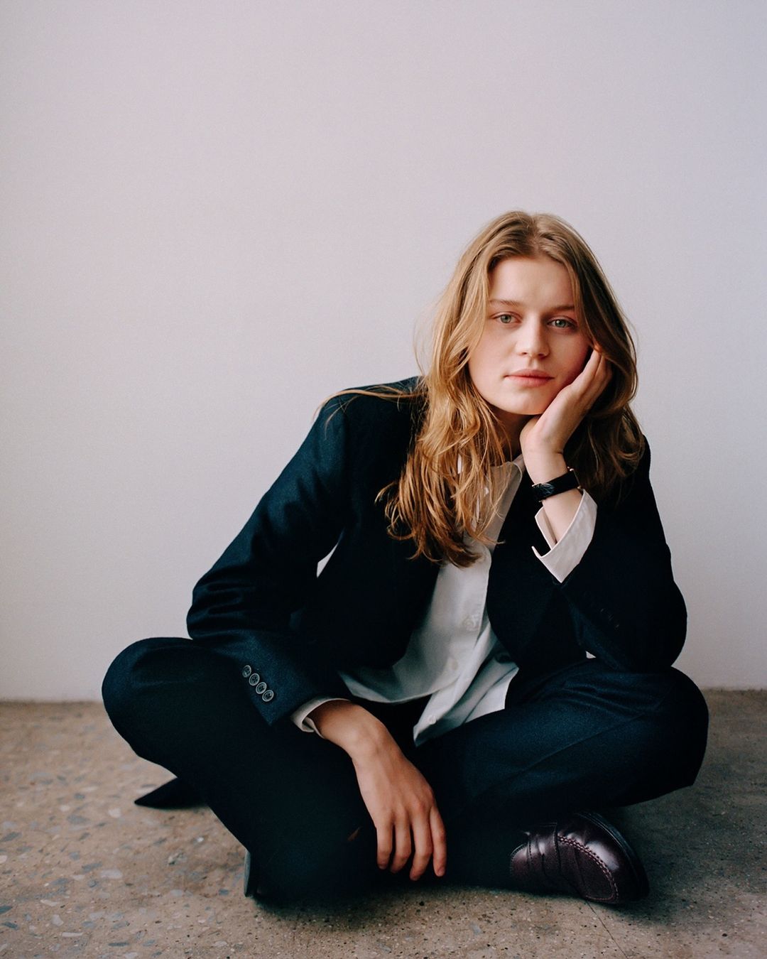 A young adult female with long blonde hair is sitting cross-legged on a concrete floor wearing a black blazer and pants in a portrait.