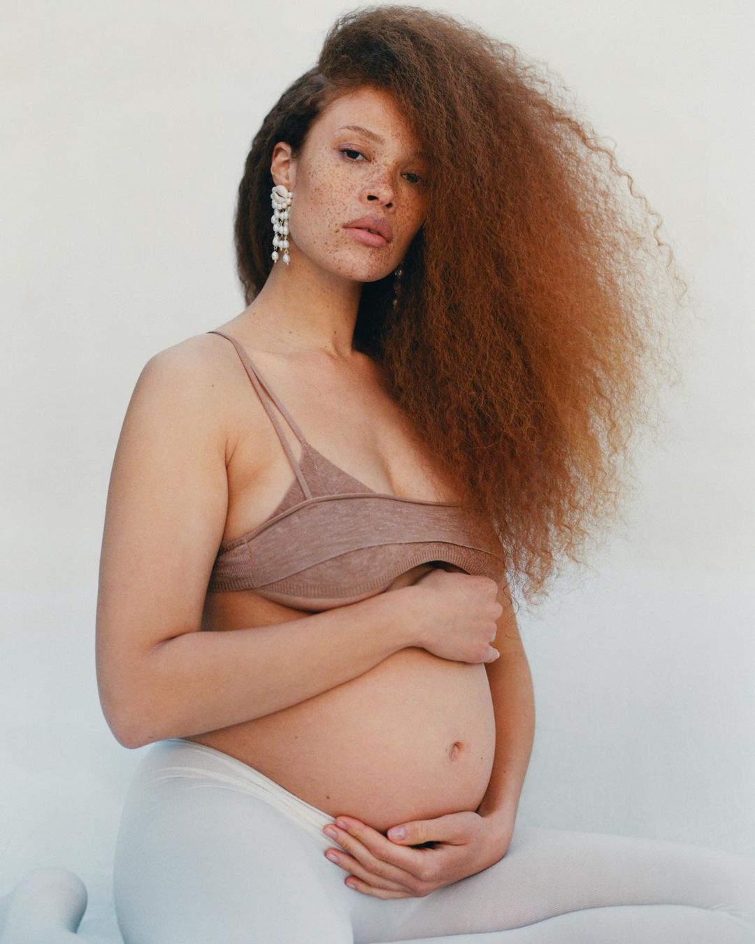 Pregnant woman with red curly hair poses in a photo studio in a neutral bra top and white tights, hands on her belly, looking at the camera.
