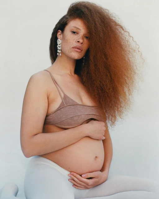 Pregnant woman with red curly hair poses in a photo studio in a neutral bra top and white tights, hands on her belly, looking at the camera.