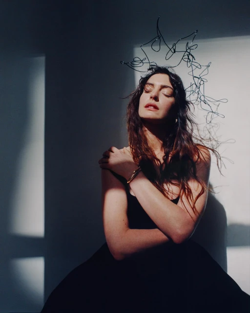 Portrait of a woman with a dark dress and an abstract headpiece posing in a studio with soft lighting and shadow casting on the white wall.