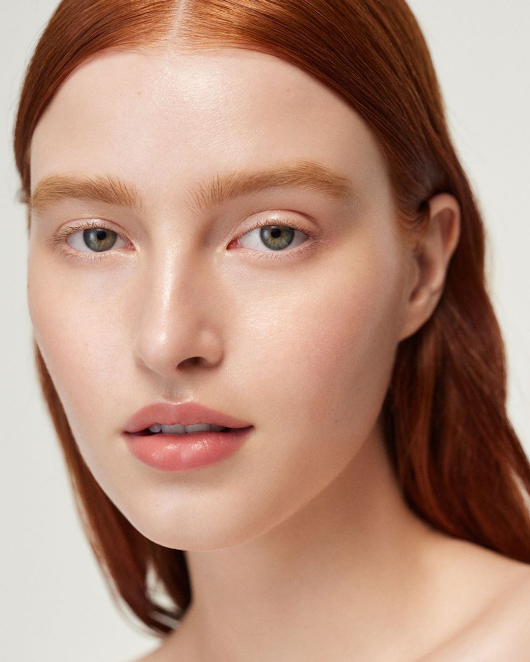 Close-up portrait of a young adult female with pale skin, red hair, and green eyes, set against a plain light background, beauty concept.