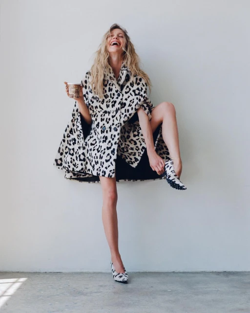 Blonde woman in animal-print coat and shoes laughs in a bright studio, holding a paper coffee cup and balancing with one leg up.