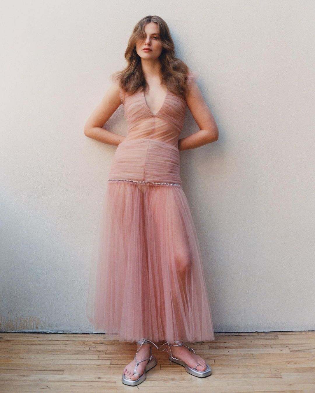 A woman in a pink gown poses against a neutral background, hands behind her. The dress is sheer with an empire waist and flowing skirt.