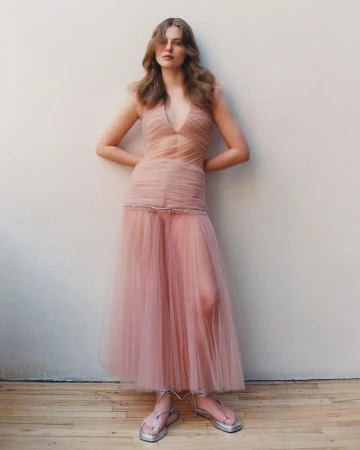 A woman in a pink gown poses against a neutral background, hands behind her. The dress is sheer with an empire waist and flowing skirt.