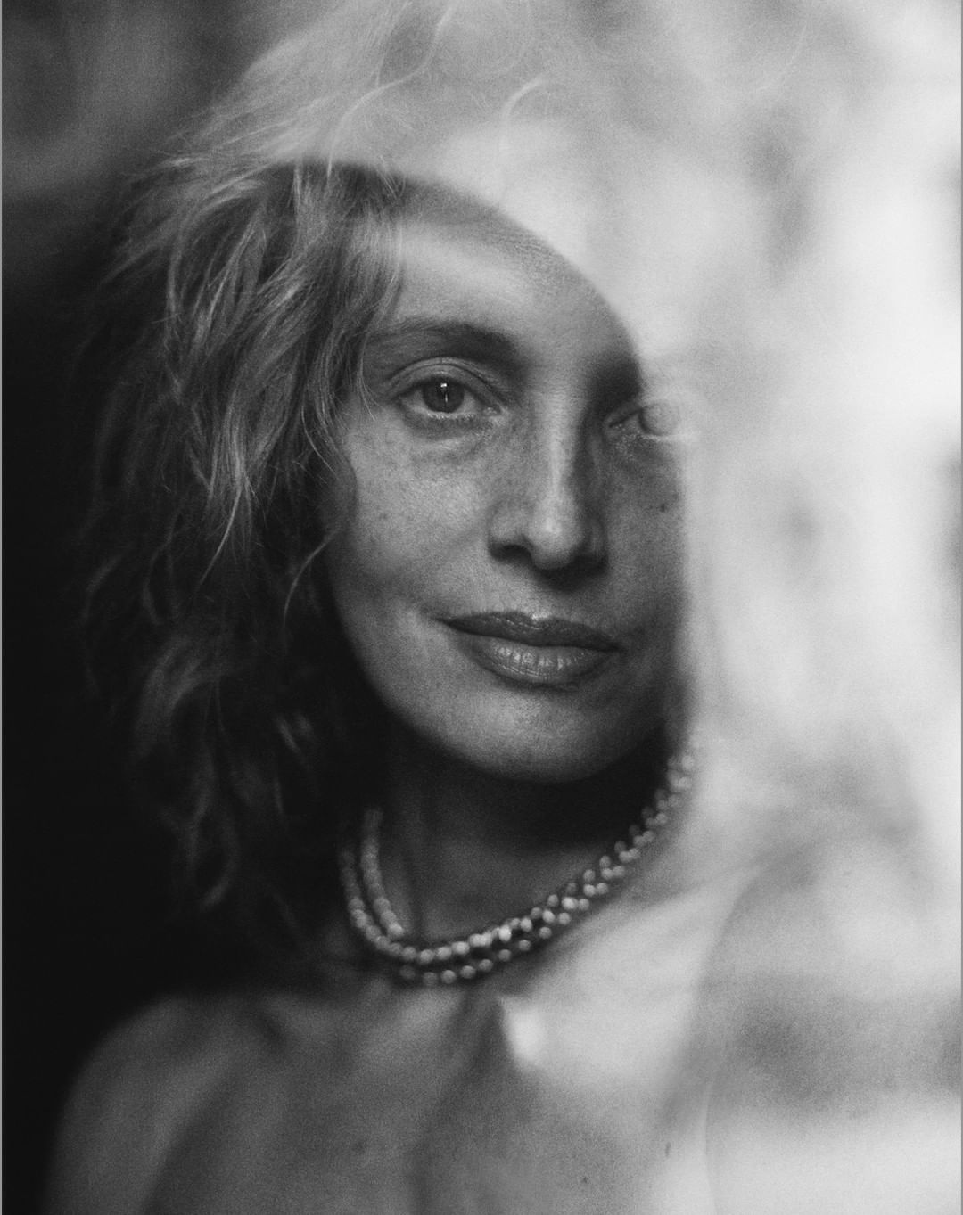 Close-up monochrome portrait of a woman wearing a double strand pearl necklace, partially obscured by light reflections.