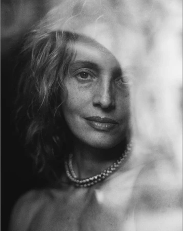Close-up monochrome portrait of a woman wearing a double strand pearl necklace, partially obscured by light reflections.