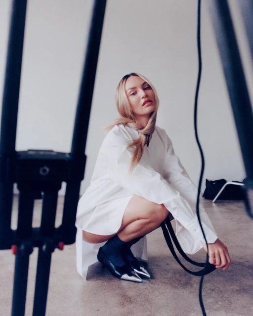 Model Candice Swanepoel poses crouching in a photo studio with a white backdrop wearing a white dress shirt and black and silver shoes.