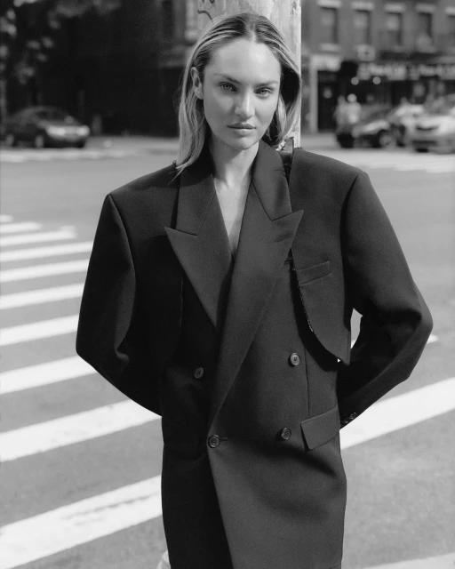 Candice Swanepoel in a black blazer dress poses on a crosswalk in an urban setting, with blurred background cars and buildings.