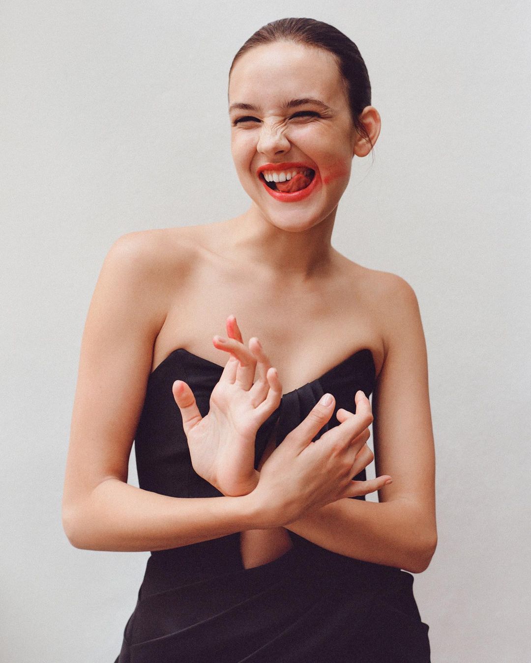 Young woman in a black strapless top with smudged red lipstick laughing against a plain white background