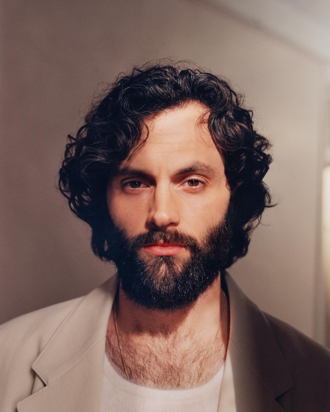Headshot of Penn Badgley.  He has dark curly hair and a full beard, wearing a cream-colored jacket. The background is a plain wall.