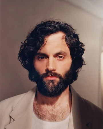 Headshot of Penn Badgley.  He has dark curly hair and a full beard, wearing a cream-colored jacket. The background is a plain wall.