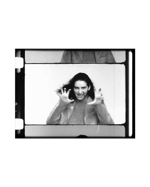 Black and white medium shot of a young adult woman with long dark hair, reaching toward the camera with an excited facial expression.