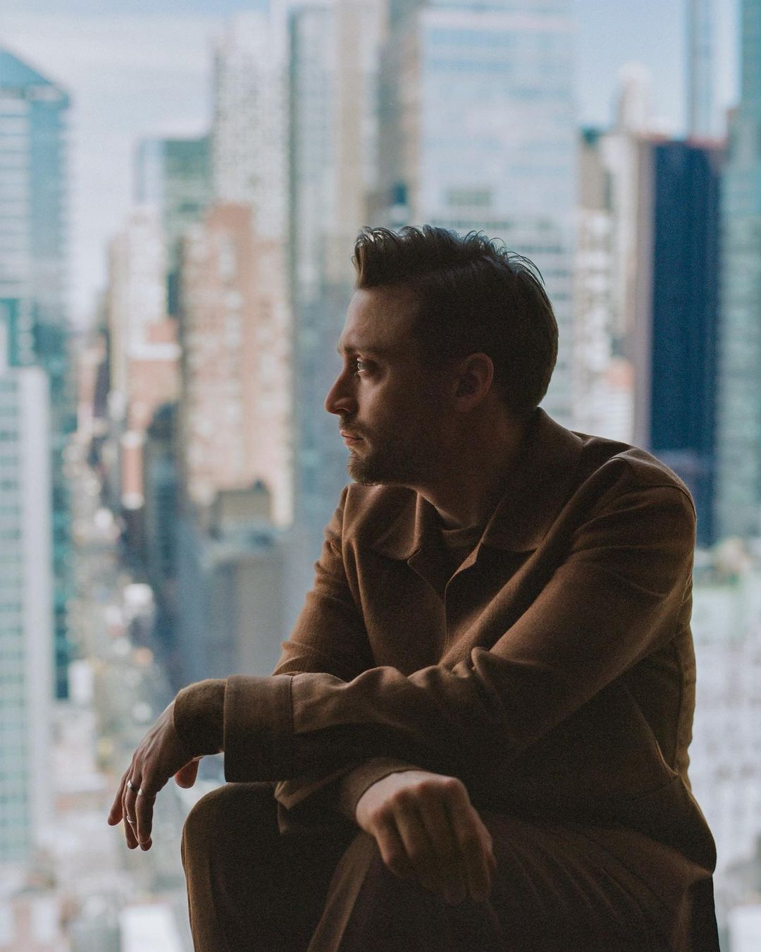 Man in brown suit sitting indoors near window with cityscape views; side view portrait with natural light.