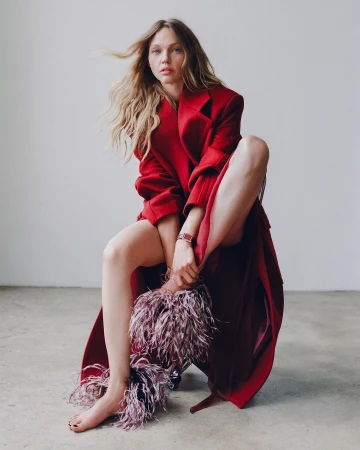 An adult woman with long blonde hair poses in a red coat. She is seated in an indoor setting with a neutral backdrop.