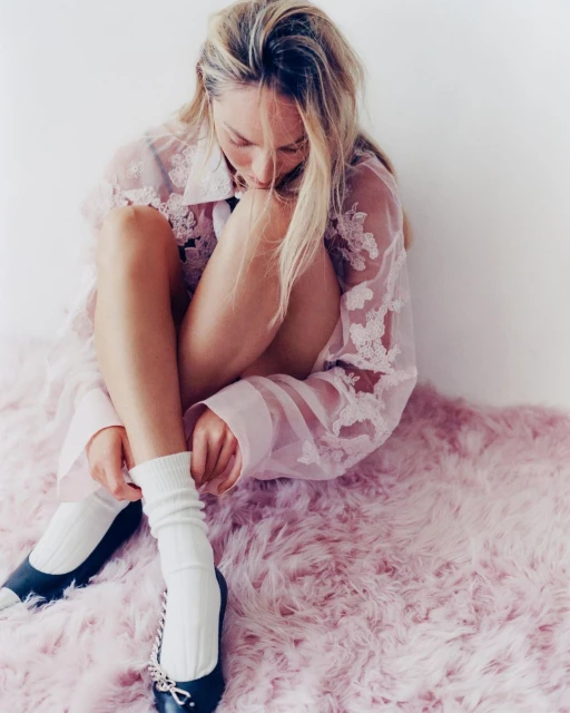 Blonde woman sits on pink fur rug putting on white socks with black ballet shoes in a soft, bright indoor fashion setting.