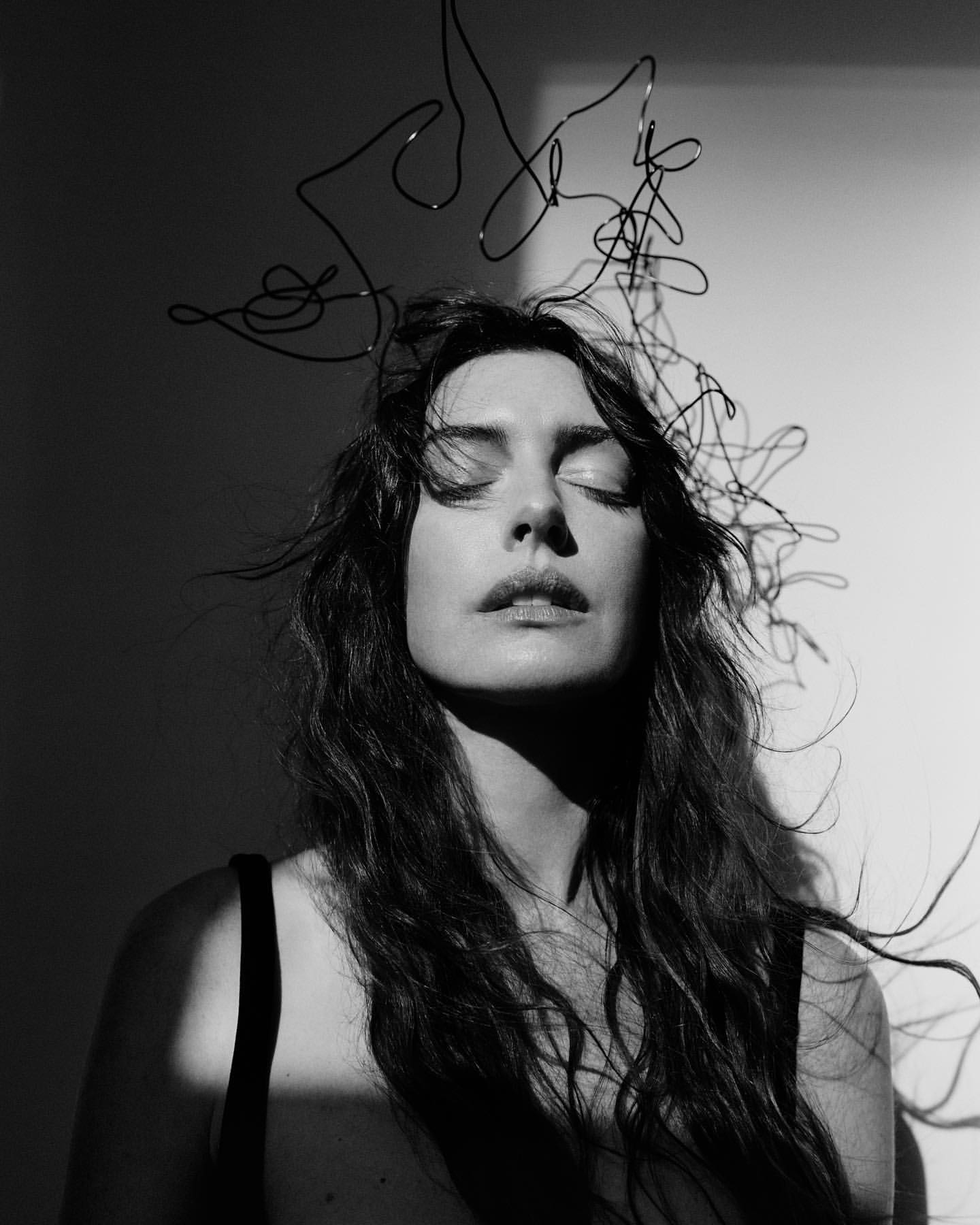 Black and white portrait of a woman with eyes closed wearing a wire headpiece against a light background with dramatic shadows.
