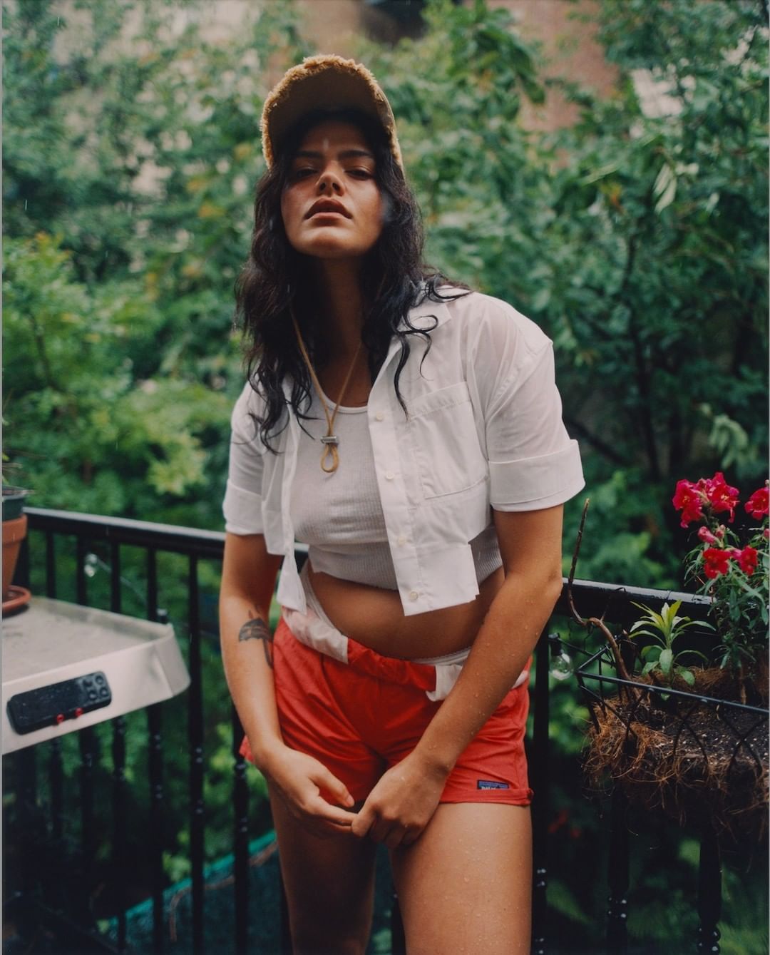 Woman with tanned skin and dark hair wears an unbuttoned white shirt and orange shorts, posing on a balcony with foliage behind her.
