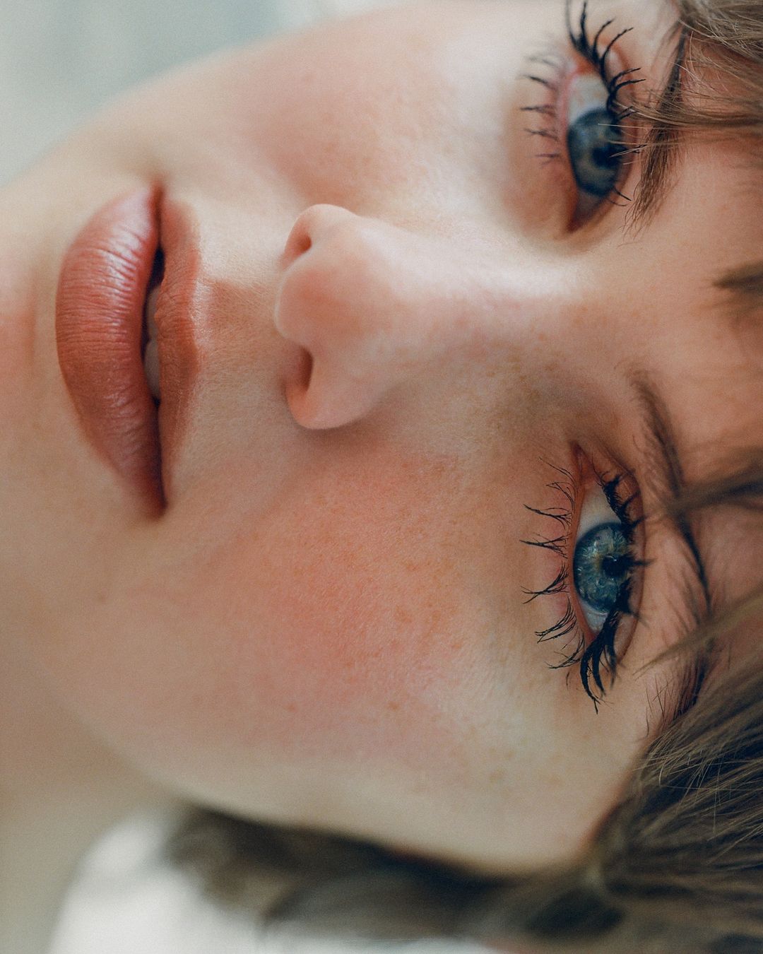 Close-up portrait of a young adult woman with blue eyes and long eyelashes, fair skin, and natural makeup, set against a blurred background.