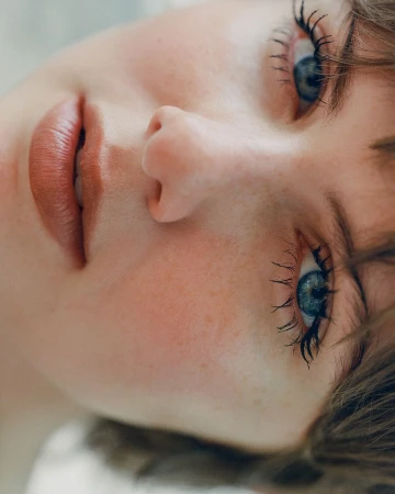 Close-up portrait of a young adult woman with blue eyes and long eyelashes, fair skin, and natural makeup, set against a blurred background.
