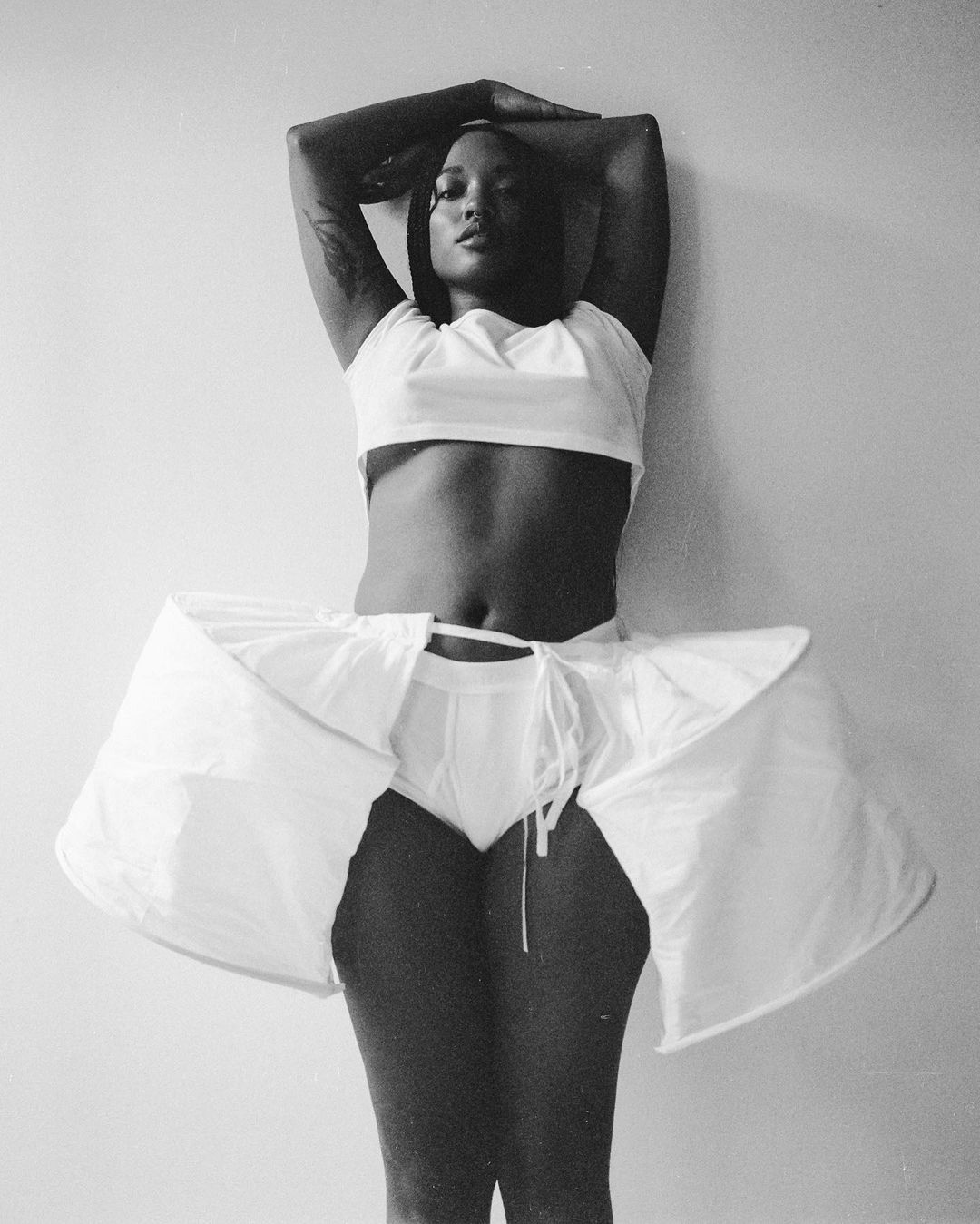A dark-skinned adult female posing indoors, wearing a white crop top, white shorts, and white crinoline, against a plain white wall.