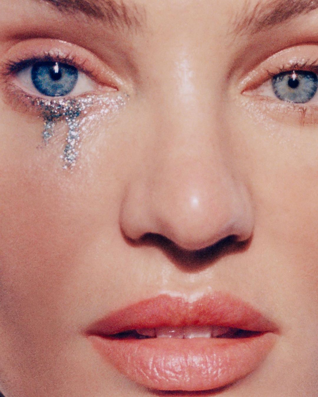 Close-up portrait of a woman's face with blue eyes, pink lips, and silver glitter tears under one eye, creating a glamorous beauty look.