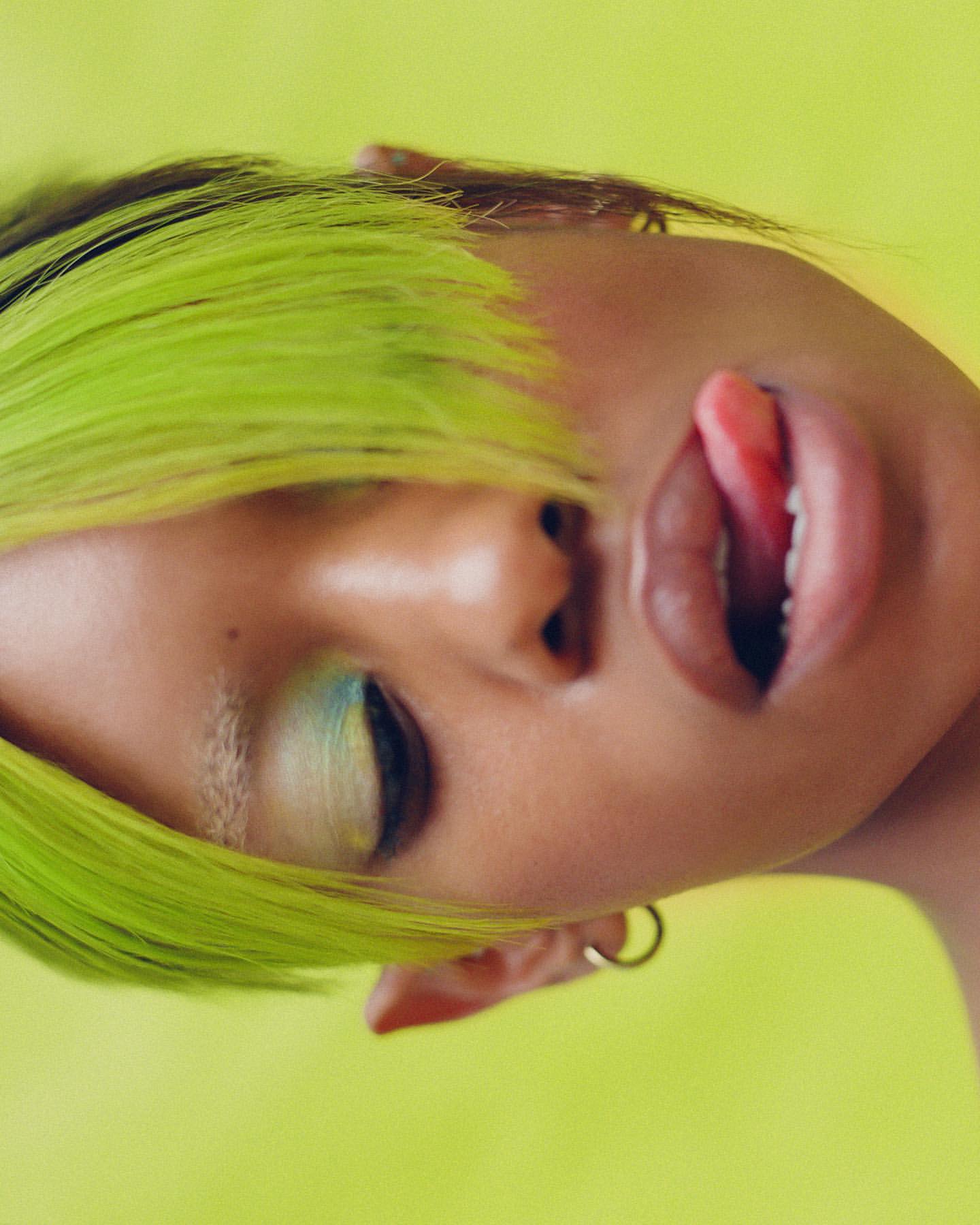 Close-up portrait of a woman with dyed neon green hair and creative blue and green eyeshadow, sticking her tongue out against a yellow background.