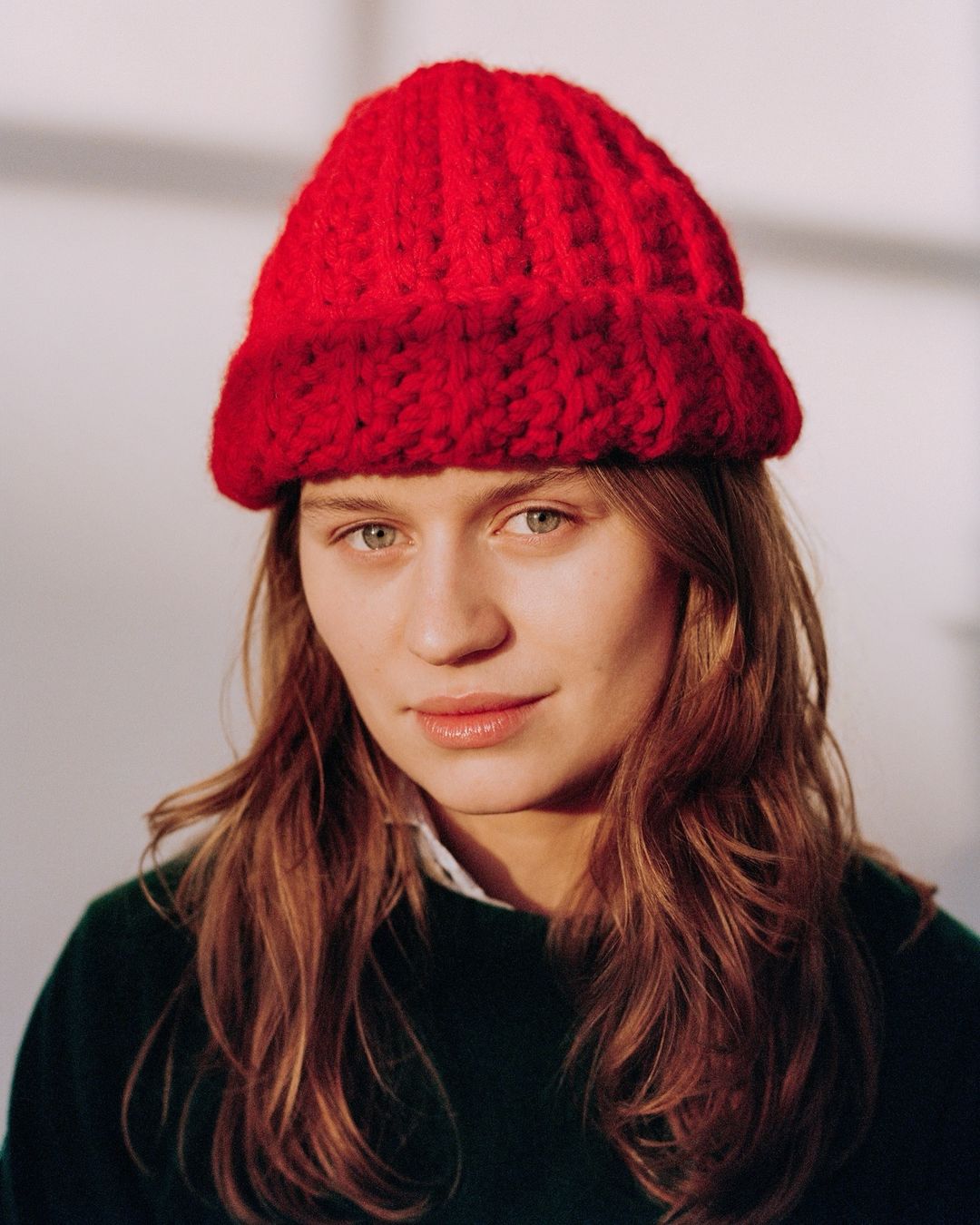 Portrait of a young woman with long wavy brown hair wearing a knitted red hat, a dark green sweater, and a white collared shirt.