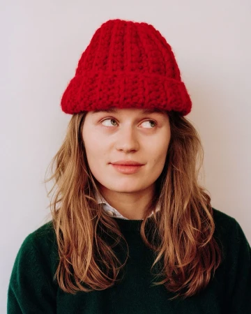 Portrait of a woman wearing a bright red knit beanie and a dark green sweater against a neutral background.