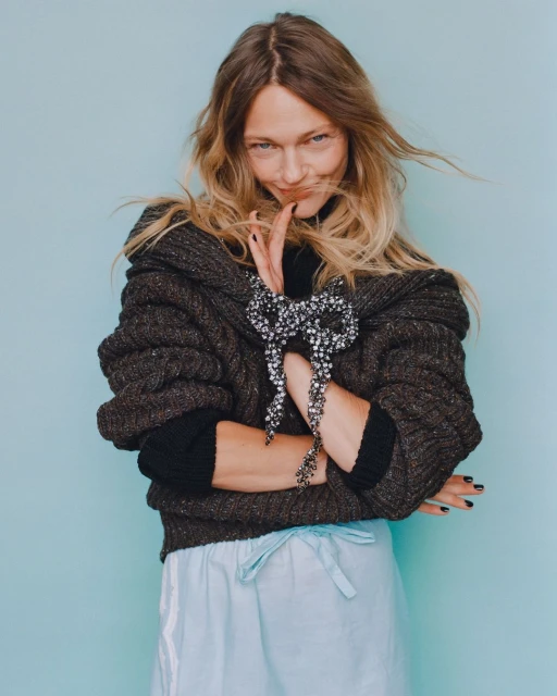 Blonde woman wearing an oversized charcoal sweater with large jewel-encrusted bow and light blue sweatpants poses on a light blue backdrop.