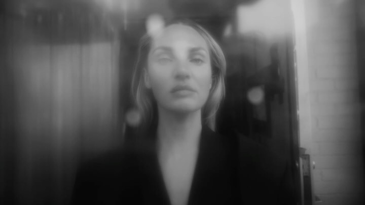 Monochrome portrait of a blonde woman with fair skin wearing a black jacket, standing indoors with diffused, soft lighting.