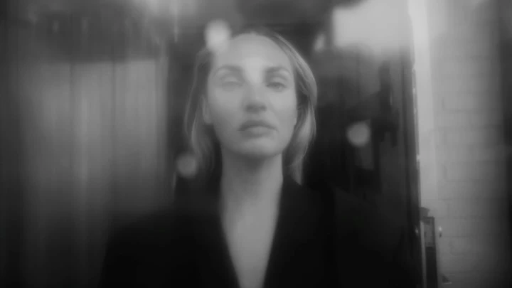 Monochrome portrait of a blonde woman with fair skin wearing a black jacket, standing indoors with diffused, soft lighting.