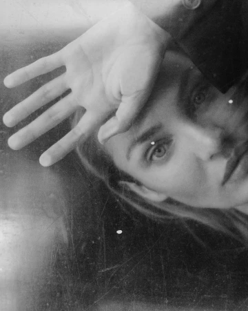 Close-up portrait of a woman with her hand raised to her forehead, shot in monochrome with textured analog grain.