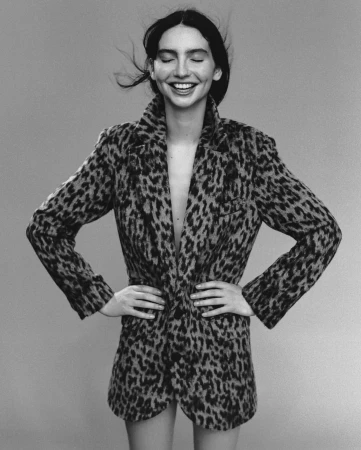A woman in a cheetah-print blazer smiles with her eyes closed, hands on hips, showcasing fashion in a studio shot with a neutral backdrop.