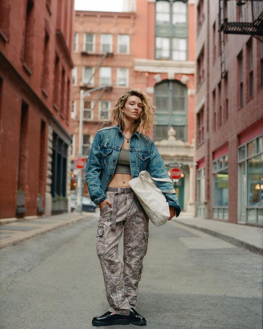 Woman in denim jacket and camo pants stands in city street. She carries a tote bag. Brick buildings line the sides of the road.