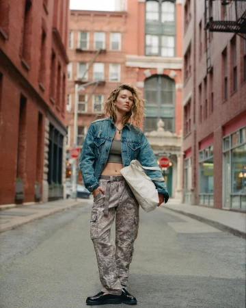 Woman in denim jacket and camo pants stands in city street. She carries a tote bag. Brick buildings line the sides of the road.