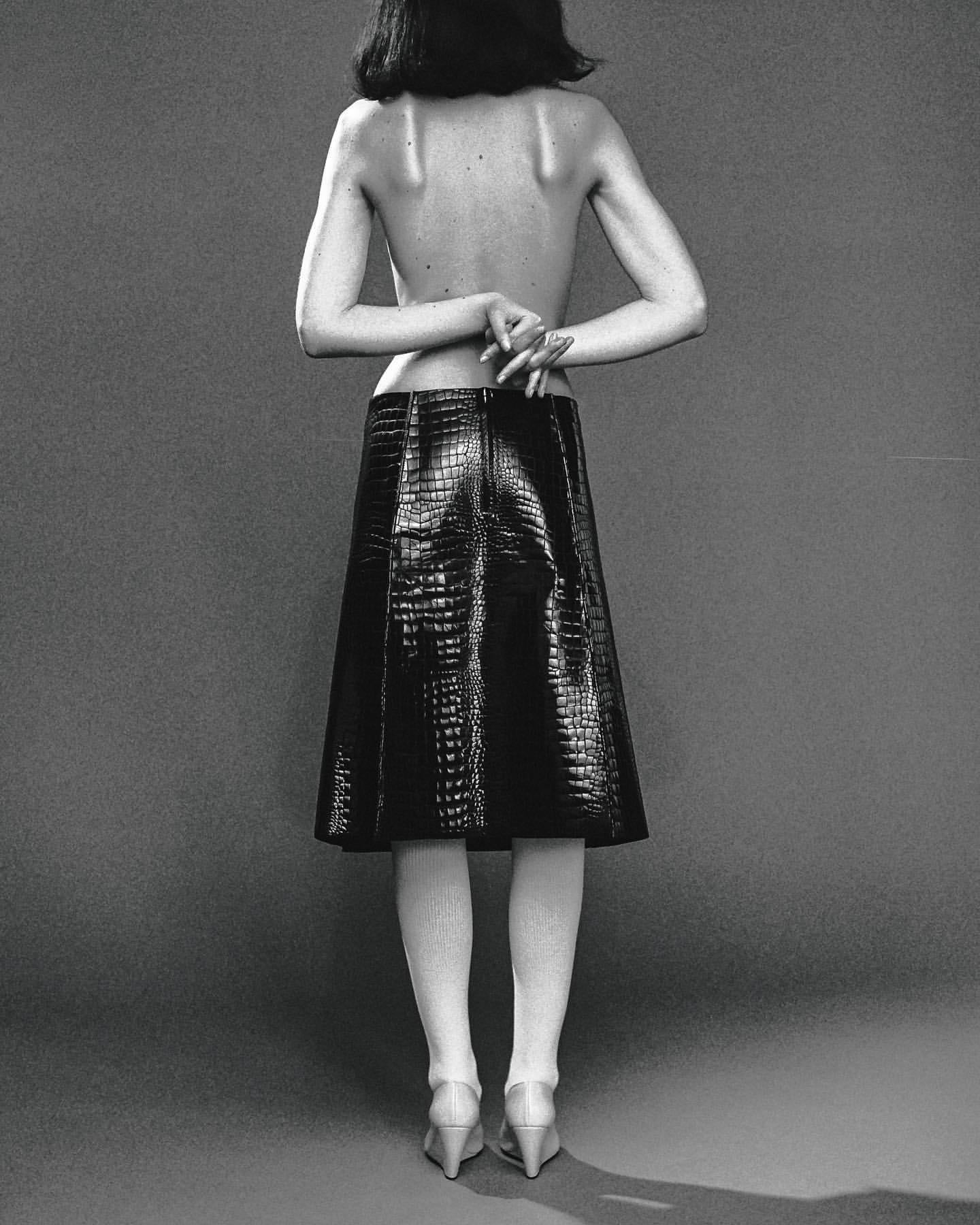 Back view of a woman with bare upper back wearing a black faux crocodile skirt, stockings, and heels, against a gray background.