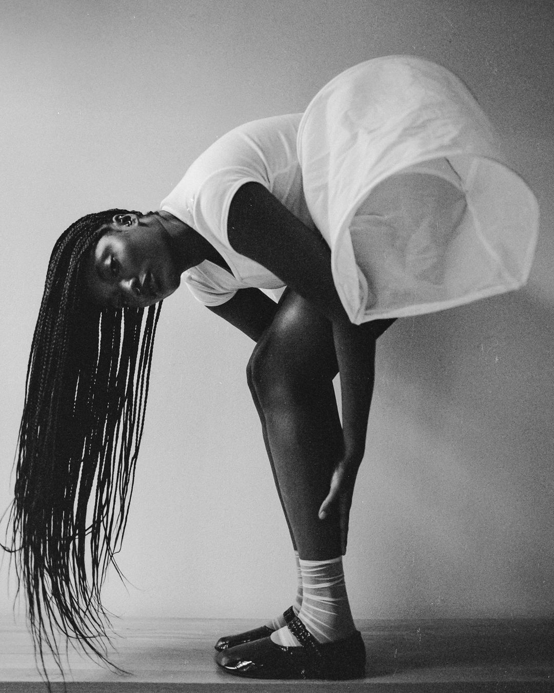 Monochromatic portrait of a Black woman bending over, hair flowing, wearing a white dress and Mary Jane shoes, with a light background.