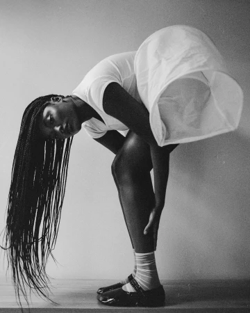 Monochromatic portrait of a Black woman bending over, hair flowing, wearing a white dress and Mary Jane shoes, with a light background.