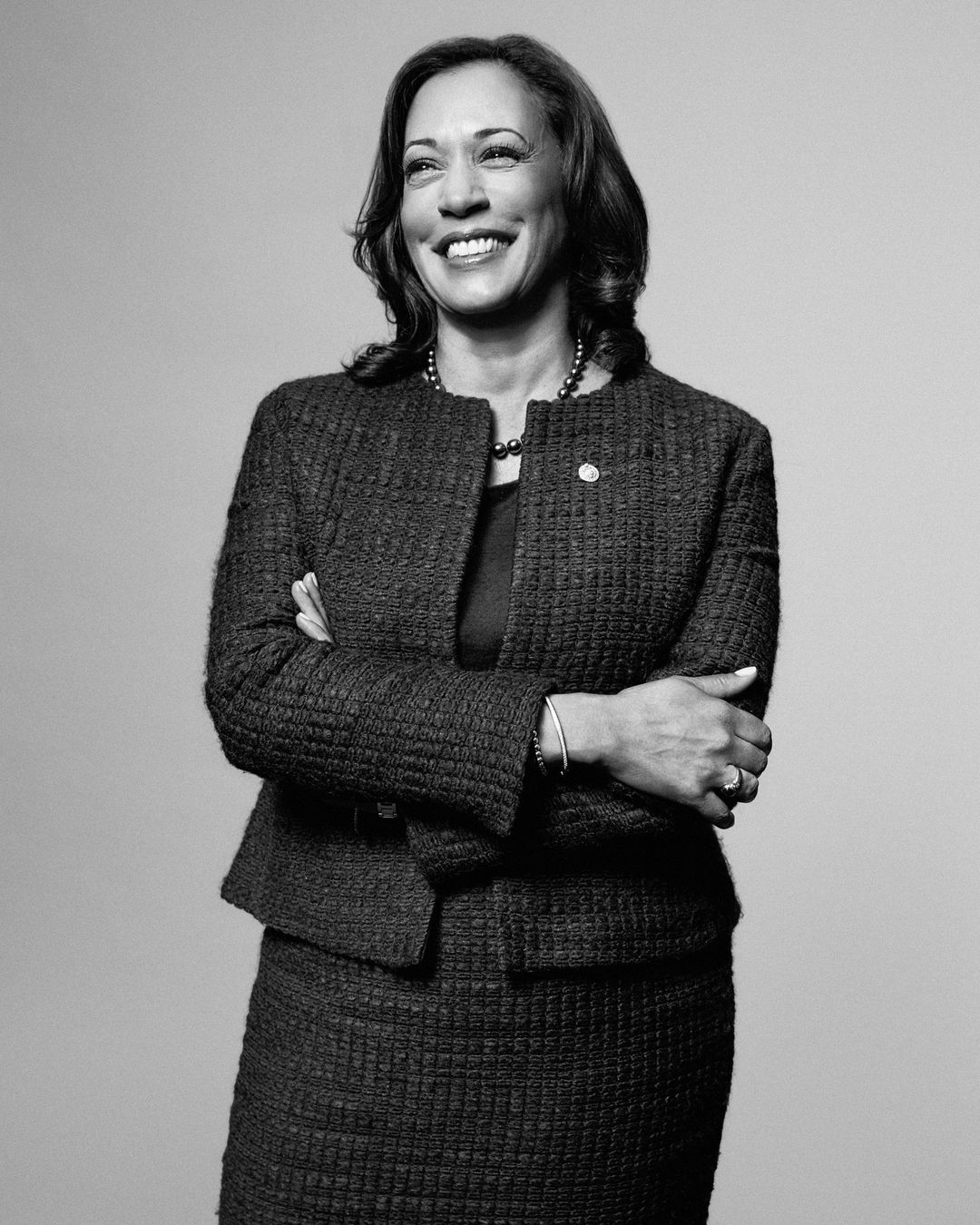 Portrait of Kamala Harris smiling with arms crossed, wearing a textured tweed jacket and skirt against a seamless grey background.