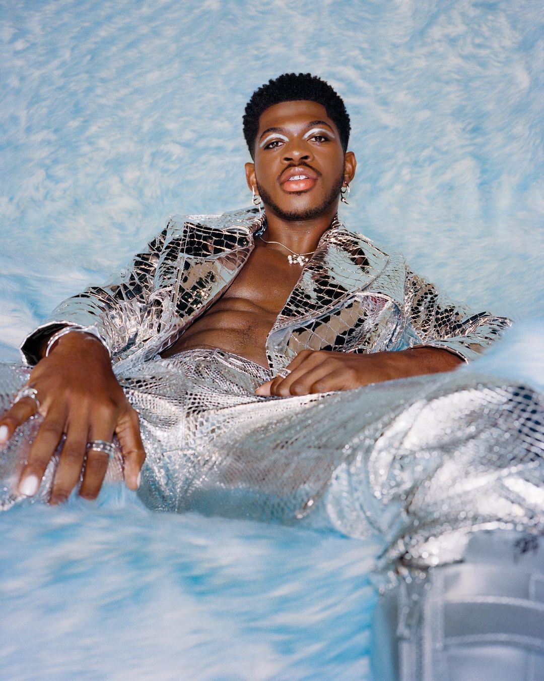 Portrait of a fashionable adult male wearing a silver jacket and pants with a patterned background. He wears jewelry and makeup.