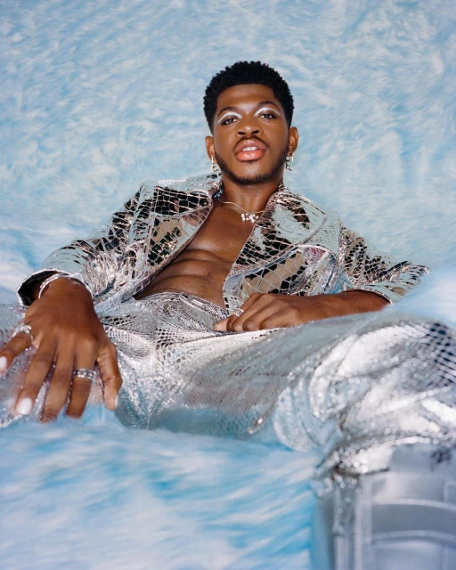 Portrait of a fashionable adult male wearing a silver jacket and pants with a patterned background. He wears jewelry and makeup.
