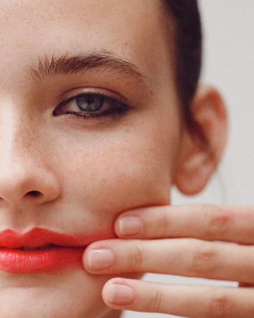 Close-up portrait of a woman with freckles, dark winged eyeliner, and red lipstick, with fingers gently touching her cheek.