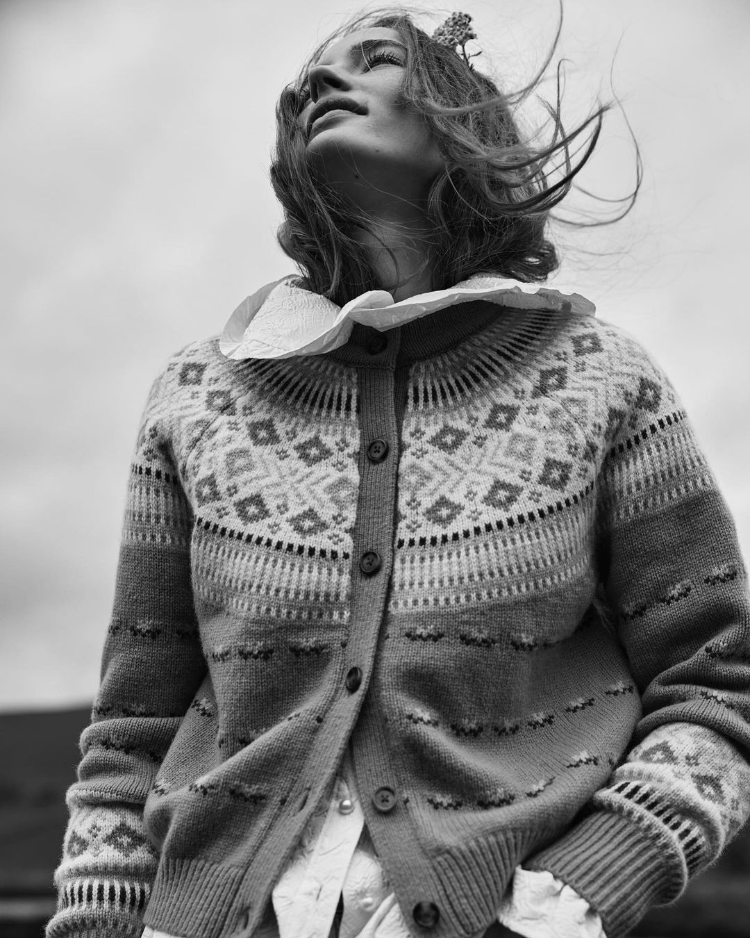 Woman in a fair isle cardigan with a ruffled collar stands outside. The wind blows in her hair against a cloudy gray sky.