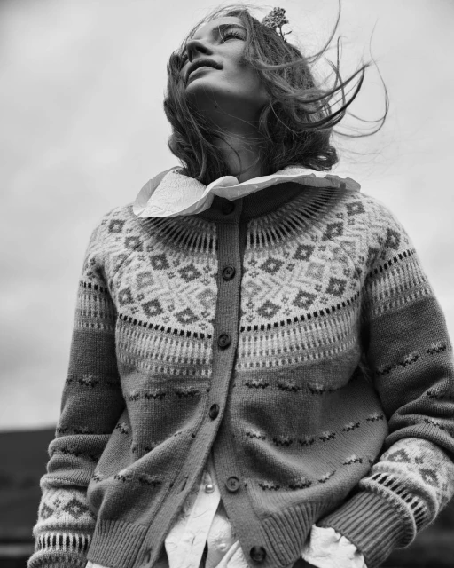 Woman in a fair isle cardigan with a ruffled collar stands outside. The wind blows in her hair against a cloudy gray sky.