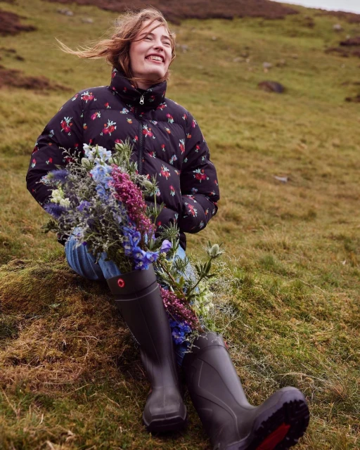 Smiling woman in floral puffer jacket and green boots sits on grassy hillside holding bouquet of purple and blue wildflowers.